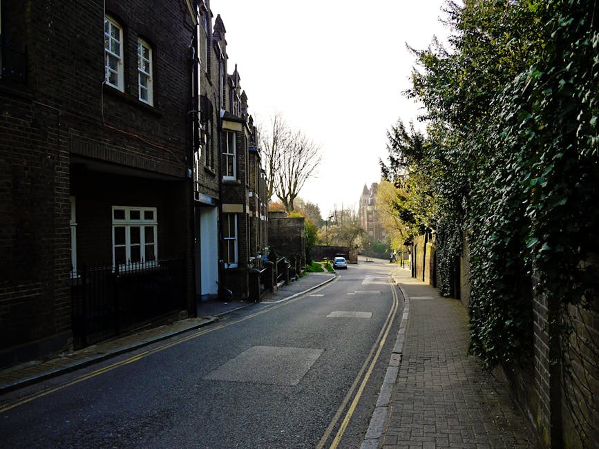 A quiet residential street in Arnos Grove during daytime, featuring a straight asphalt road bordered by well-maintained brick and wooden fences, with neatly trimmed hedges and lush green trees lining