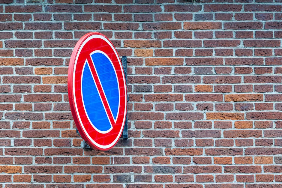 A round traffic sign is mounted on a brick wall outside a property, indicating no parking or stopping in the area. The sign features a red border with a blue background and a red diagonal line across the center, representing prohibition. The brick wall behind the sign is constructed of reddish-brown bricks with visible mortar lines, and the surface appears clean and well-maintained. In a house removal or home relocation context, such signage may be relevant for understanding parking restrictions during moving days. The setting is outdoors, with natural lighting illuminating the scene. Occasionally, Man with Van Osidge's removals service might manage vehicle parking and logistics within regulated areas, ensuring compliance with local rules like those indicated by the sign, especially when performing furniture transport or packing and moving activities near residential properties in Osidge. The image does not depict any moving equipment, furniture, or personnel directly involved in the relocation process.