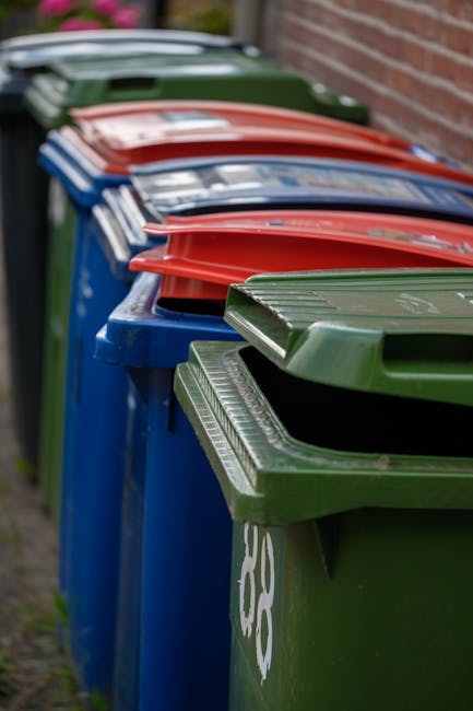A row of blue, green, and red wheelie bins with closed lids, positioned outside a residential building along a paved pathway beside a brick wall. The bins are used for waste disposal and recycling, and some have labels or symbols indicating the type of waste. The background shows the edge of a garden or yard with some greenery. The image captures the bins during daylight, with natural light illuminating the scene. This scene relates to household waste management as part of home relocation or clearance activities, and is associated with professional removals services such as those provided by Man with Van Osidge, who assist with house and garden waste disposal as part of move logistics.