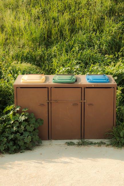 Three large wheeled trash bins, one green and two black, are positioned side by side on a paved driveway in front of a tall, dense hedge of green foliage. The bins are placed near the edge of the property, with their lids closed, indicating they are awaiting collection or disposal. The surface of the driveway consists of light-colored stones or concrete paving, and the scene appears to be outdoors during daylight hours. This setup could be part of a house removal or garden clearance process handled by professional services such as Man with Van Osidge, which offers home relocation and waste disposal solutions relevant to the page about large garden waste and shed disposal options in Osidge.