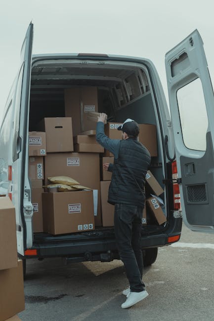 A person wearing a dark jacket, black pants, white sneakers, and a black cap is standing at the rear of an open white moving van, which is parked outside a property. The individual is reaching inside