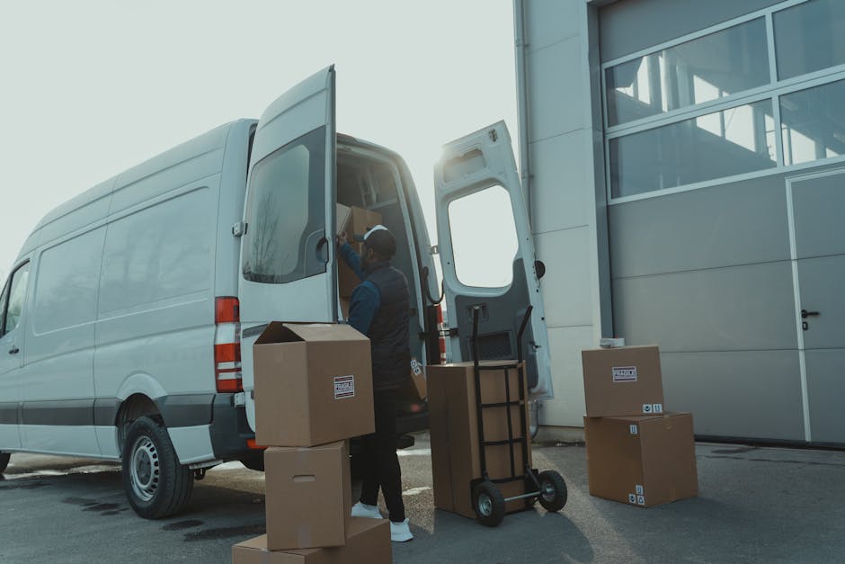 A person wearing a dark jacket, black pants, white sneakers, and a black cap is standing at the rear of an open white moving van, which is parked outside a property. The individual is reaching inside