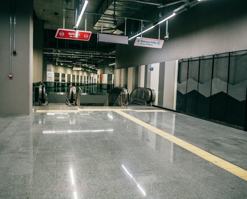 The image depicts an underground metro station entrance with a smooth, polished grey floor reflecting the overhead lighting. There are two escalators in the background, one going up and one going down, enclosed by black safety barriers. To the right of the escalators, there's a wall with a modern geometric black and grey design. Hanging overhead are signs indicating directions in English and another language, including a red sign that reads 'Girilmez No Entry' and a white sign pointing towards 'Metroya Gidiş.' The station is vacant, with no people visible, and the environment is illuminated by bright, white LED lighting attached to the exposed ceiling. The space appears clean and well-maintained, typical of a city transport hub. Such environments are commonly encountered during home relocation projects that involve furniture transport or packing and moving services, as provided by companies like Man with Van Osidge.