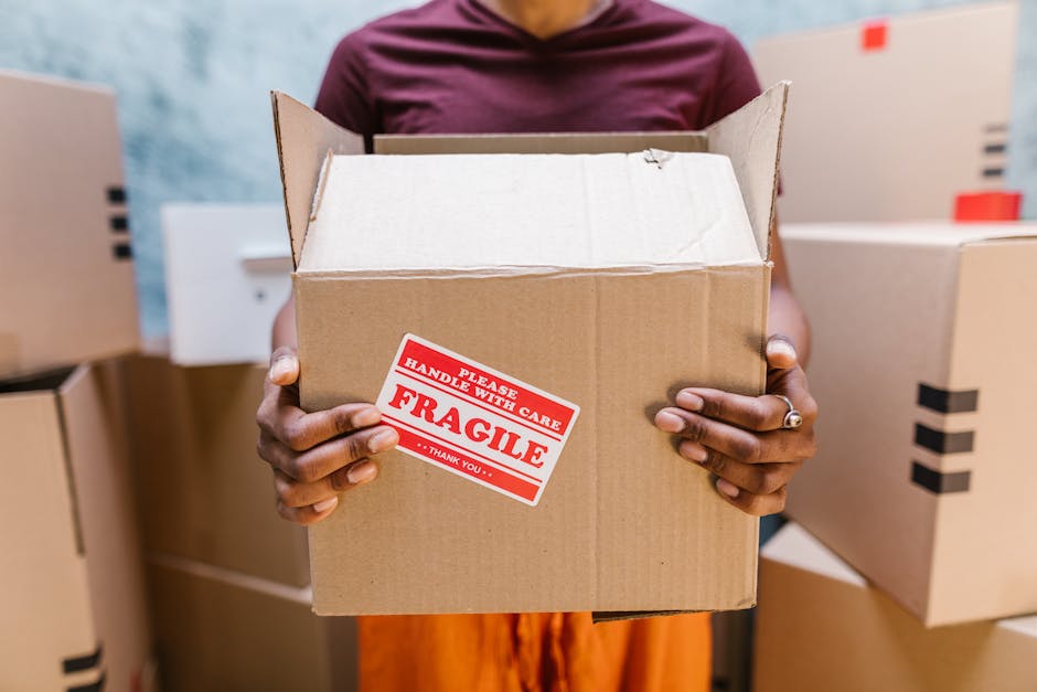 A man and a woman stand inside a doorway at an apartment entrance, engaged in home relocation tasks. The man, dressed in a maroon shirt and loose-fitting orange trousers, holds a large cardboard box w