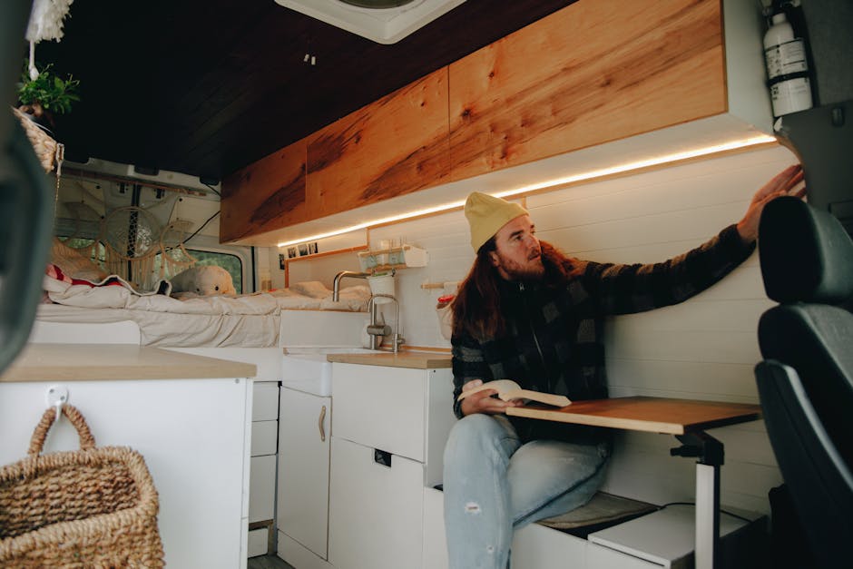 Inside a compact, modern vehicle used for home relocation, a young man with long hair, a beard, wearing a beige beanie, a black and gray checkered shirt, and light jeans, is seated at a foldable table attached to the side wall of the van. He is holding a book in his left hand and reaching out with his right hand to adjust or secure a panel on the white interior wall of the vehicle, which features horizontal paneling. The vehicle's interior includes shelves, small cabinets, and visible storage spaces, with soft warm lighting embedded beneath a wooden overhead storage compartment made of natural wood. To the left, there is a woven basket in the foreground, and a bed or mattress with plush toys and soft bedding is seen towards the back, indicating that the van is being used for moving personal belongings and furniture. The environment appears organized and designed for efficient packing and furniture transport, with the focus on the interior space prepared for the loading process, aligning with house removal and logistical services by Man with Van Osidge.