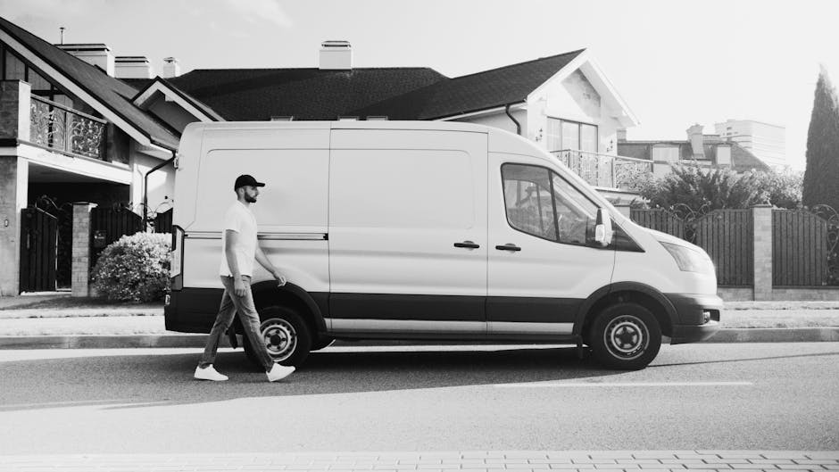 A white USPS delivery van parked on a residential street with multiple cars lined up alongside. The van displays the USPS logo and a 'Now Hiring' sign on its rear door, along with a website address. T