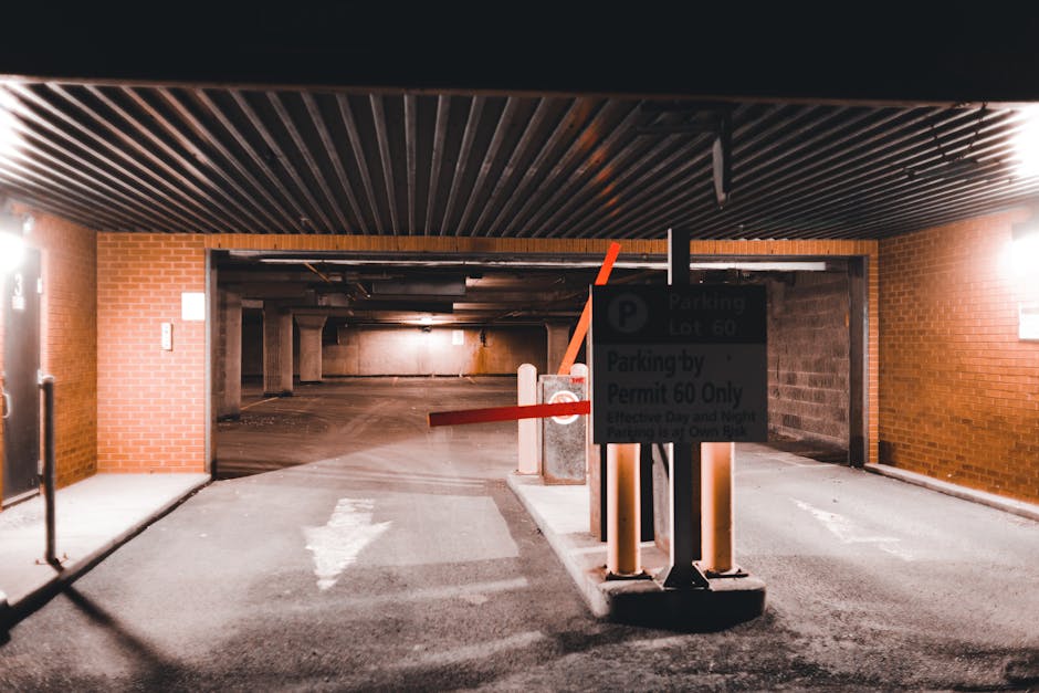 The image shows an indoor parking garage with a brick wall on both sides and a black ceiling with parallel slats. At the entrance to a designated parking area, there is a sign indicating parking lot 60 and restrictions for parking by permit only. In front of the entrance, there are two vertical white bollards connected by a red horizontal barrier arm that is partially lowered, preventing entry. The ground is paved with asphalt, and a white arrow painted on the pavement indicates the direction of travel into the lot. Inside the parking garage, ambient lighting illuminates the space, revealing concrete pillars supporting the structure and a ramp leading downward into the parking area. The scene suggests a location where a professional removals service, such as Man with Van Osidge, might be involved in transporting household items or furniture during a home relocation, with the parking permits and bollards in place to manage vehicle access for moving vehicles.