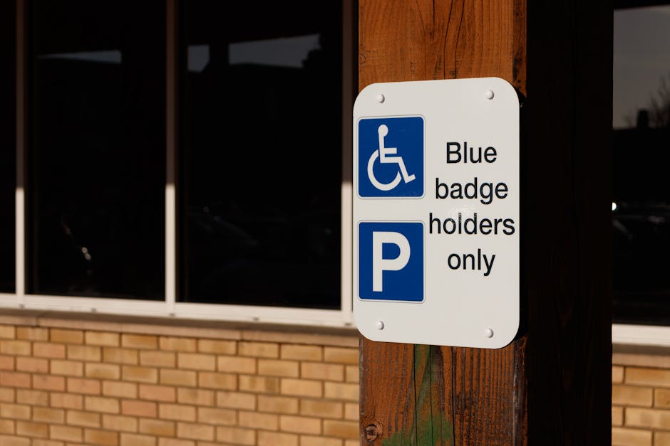 A white rectangular parking sign attached to a wooden post outside a building featuring the blue wheelchair symbol with the text 'Blue badge holders only' and a large blue 'P' symbol beneath it. The sign indicates designated parking for blue badge holders, with the background showing a brick wall and large glass windows of the building. The area appears to be an accessible parking space, relevant to house removals and relocating services by Man with Van Osidge, emphasizing logistical considerations during home relocation or furniture transport. The image is well-lit, capturing the sign's details and its surroundings clearly, reflecting parking arrangements that may facilitate the loading and unloading process during furniture packing and moving.