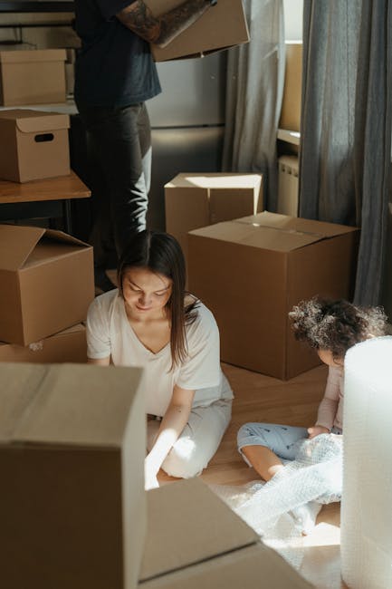 A woman and a young girl are inside a room during a home relocation process, surrounded by several large cardboard boxes, some open and some sealed, indicating packing and moving activity. The woman,