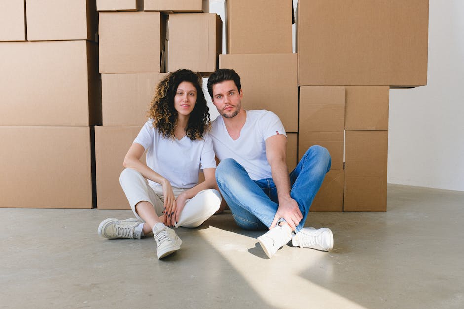 A woman and a young girl are inside a room during a home relocation process, surrounded by several large cardboard boxes, some open and some sealed, indicating packing and moving activity. The woman,