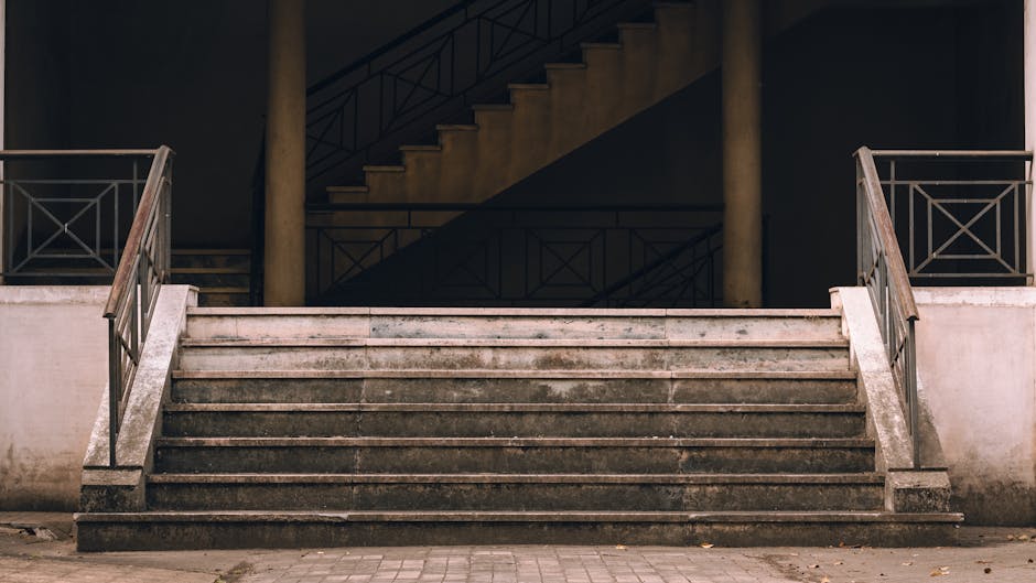 A set of wide, weathered stone stairs leading up to a building with a covered walkway featuring dark metal railings and support columns. Behind the stairs, a staircase with dark steps and matching railings ascends to an upper level, partially visible against a dark background. The space is illuminated by ambient lighting, highlighting the textures of the stone and metal. This scene depicts a typical home or commercial property entrance, and it is relevant to house removals and transportation services offered by Man with Van Osidge, especially when stairs pose a challenge during furniture transport or loading and unloading during a home relocation.