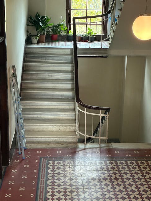 The image shows an interior staircase in a residential building with a curved wooden handrail and white metal balustrades leading up to a landing with a large window. On the landing, several potted plants are arranged on a windowsill, allowing natural light to illuminate the area. The staircase is positioned adjacent to a beige wall, with a vintage-style tiled floor featuring intricate geometric patterns in shades of red, cream, and gray. To the left of the staircase, a silver step ladder leans against the wall, indicating ongoing moving or packing preparations. A round wall-mounted light fixture emits a warm glow on the right side of the image. The scene captures a moment typical of home relocation or furniture transport, where careful handling of interior features is essential during removals, with [COMPANY_NAME] providing professional services to manage such logistics efficiently.