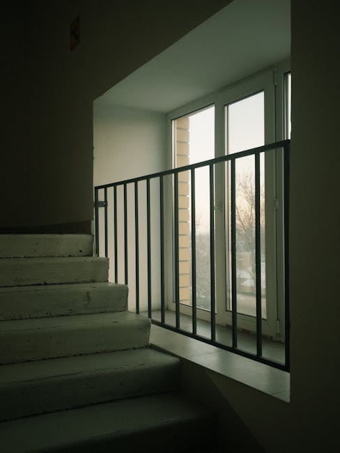 A set of wide, weathered stone stairs leading up to a building with a covered walkway featuring dark metal railings and support columns. Behind the stairs, a staircase with dark steps and matching railings ascends to an upper level, partially visible against a dark background. The space is illuminated by ambient lighting, highlighting the textures of the stone and metal. This scene depicts a typical home or commercial property entrance, and it is relevant to house removals and transportation services offered by Man with Van Osidge, especially when stairs pose a challenge during furniture transport or loading and unloading during a home relocation.